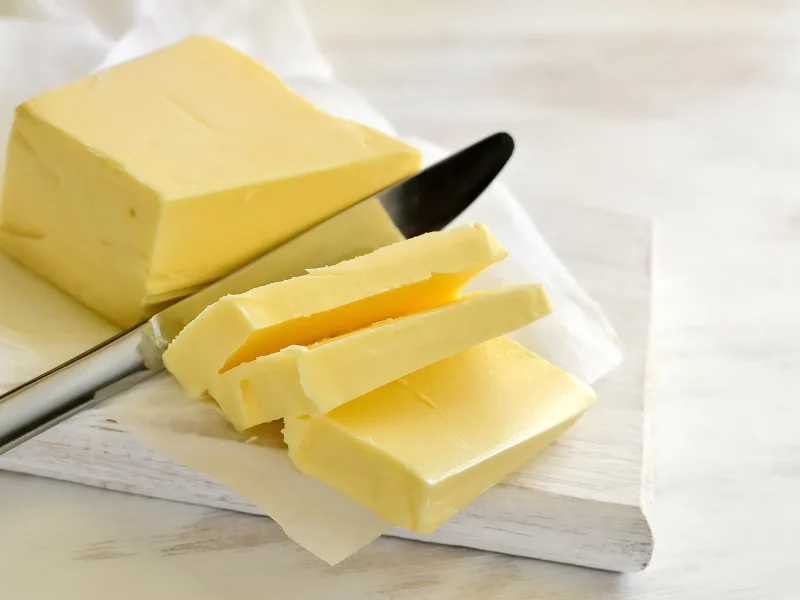 A block of fresh butter sliced with a knife, showing its creamy texture on a white wooden cutting board.