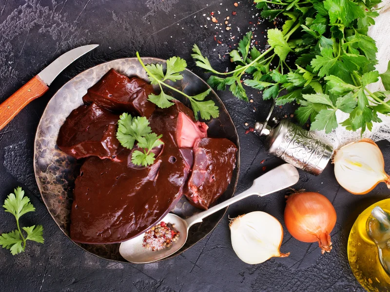 A plate of raw beef liver with fresh parsley, onions, and peppercorns, with a knife placed beside it on a rustic surface.