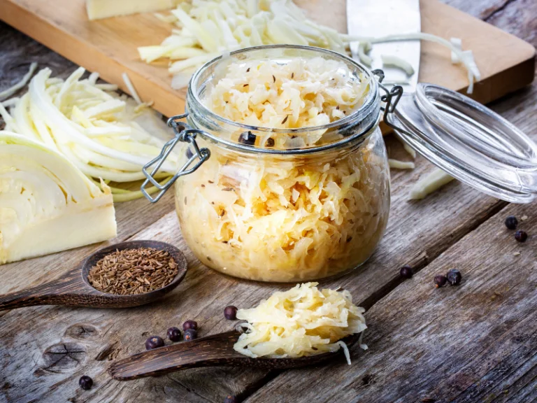 A jar of sauerkraut, rich in vitamin k2, with freshly sliced cabbage and spices on a wooden cutting board.